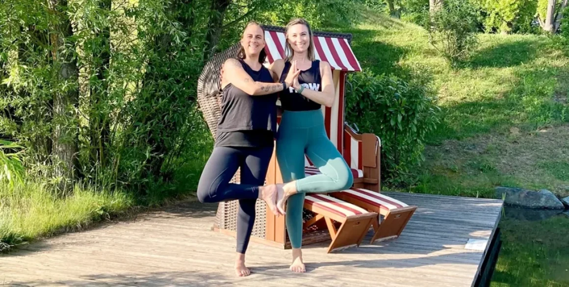 Zwei Frauen praktizieren Yoga in einer harmonischen Pose auf einem Holzdeck im Freien.
